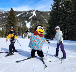 diamond peak's mascot penguin pete in a tropical shirt on the slopes with a couple of skiers smiling